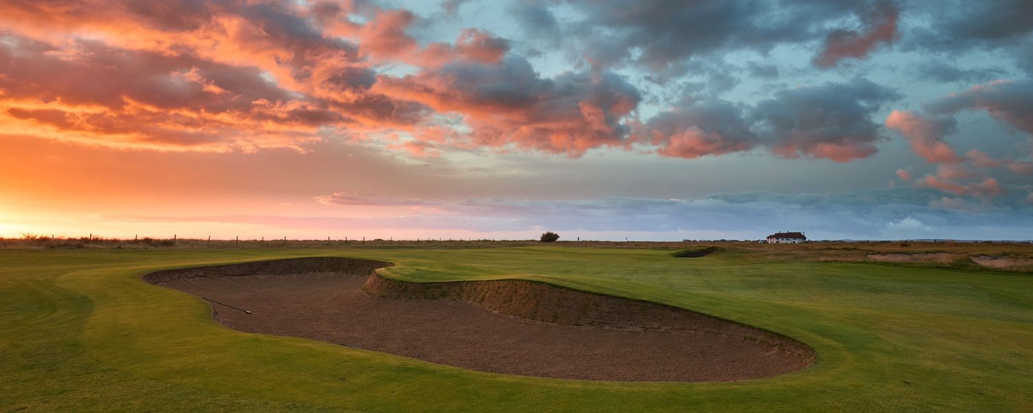 Dramatic sunset sky over a links golf course with a large sand bunker in the foreground.