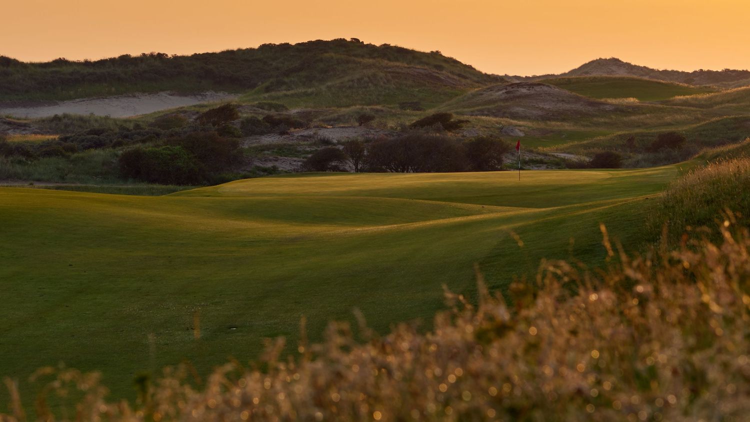 A lush golf course fairway at sunset with rolling sandy dunes and warm golden sky in the background.