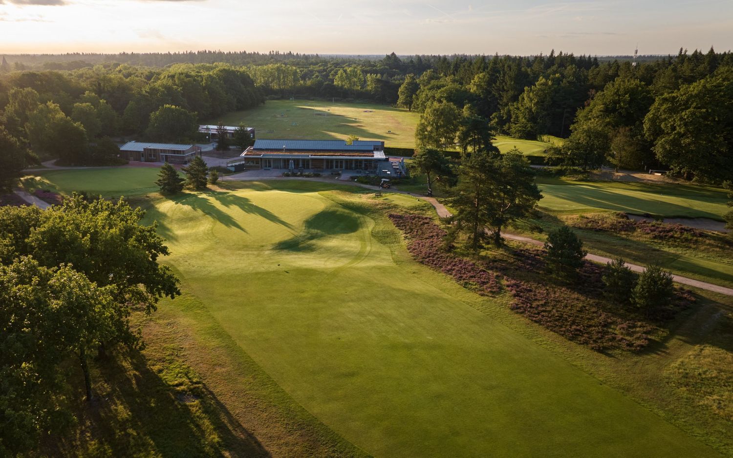 Aerial golden hour view of a parkland golf course with a clubhouse surrounded by mature trees and open fairways.