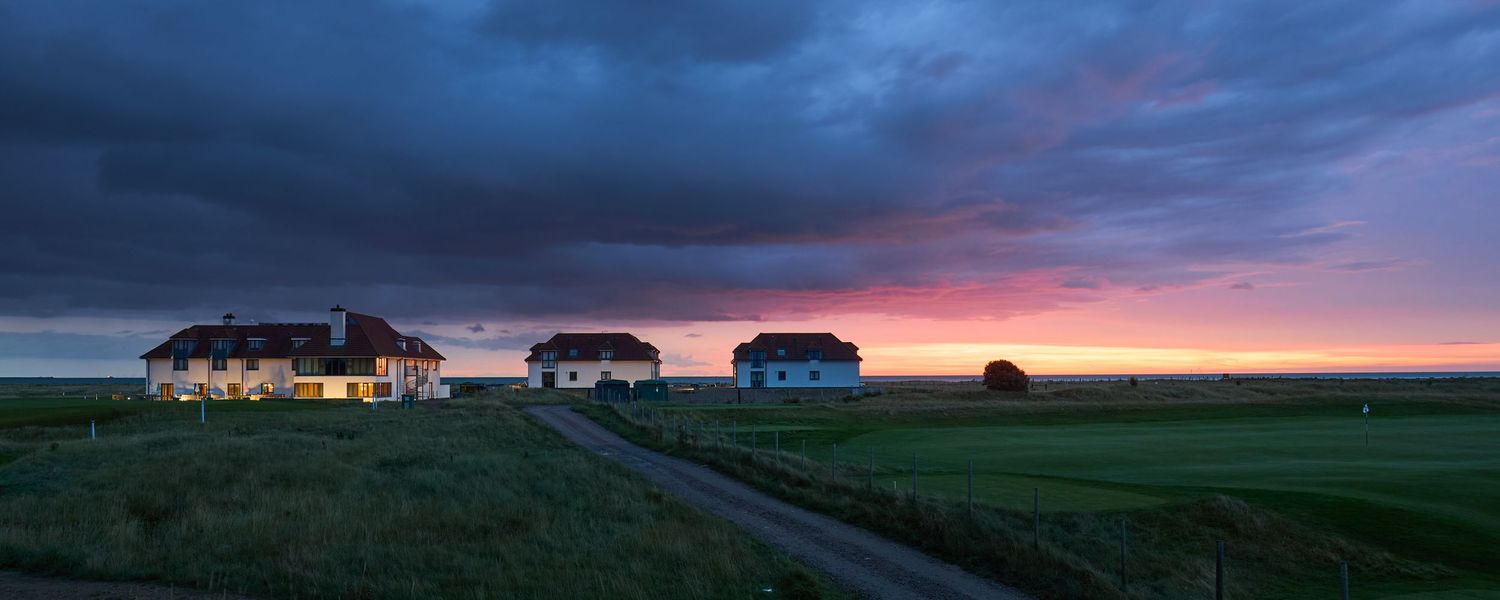 Charming golf clubhouse buildings silhouetted against a dramatic pink and purple sunset sky.