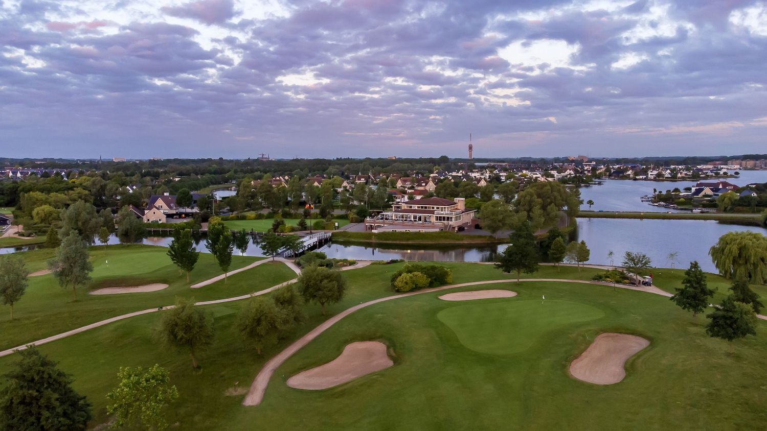Aerial view of a golf course with sand bunkers beside a scenic lake and town skyline.