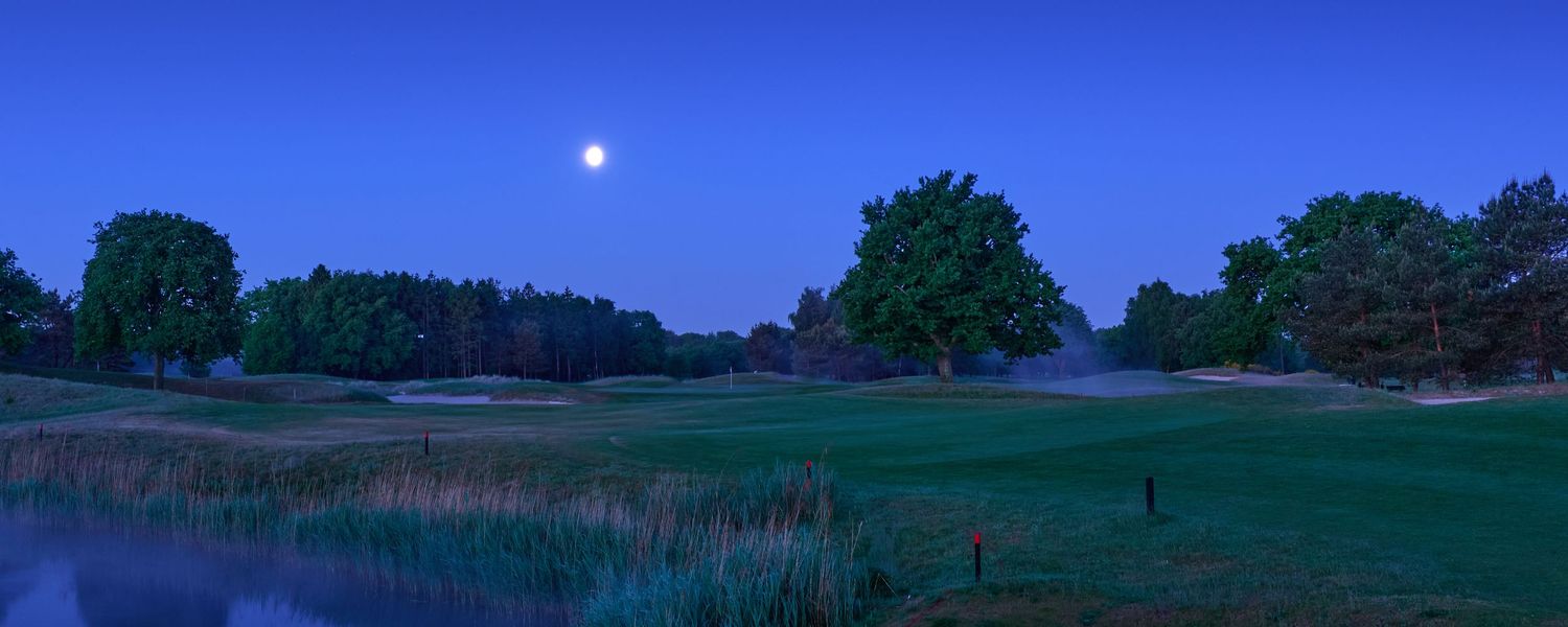 Golf course at dusk with full moon rising over lush green fairways and tranquil pond reflecting twilight sky.