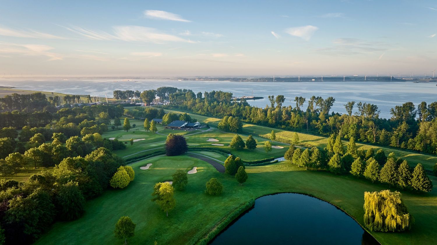 Aerial view of a golf course with water features and lush greens beside a misty lake.