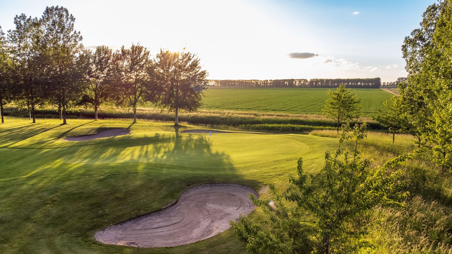 Golden hour view of a golf course green with a large sand bunker surrounded by trees.