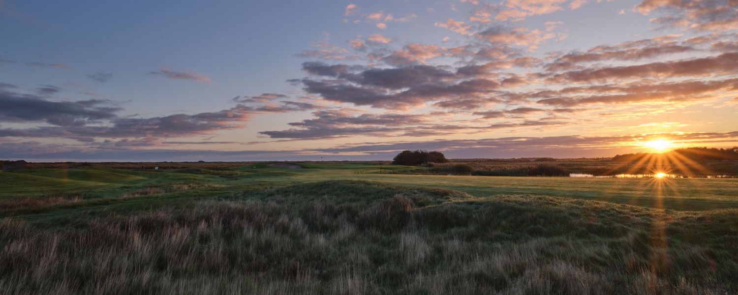 Panoramic sunrise view over a links golf course with dramatic clouds and golden sunburst on horizon.