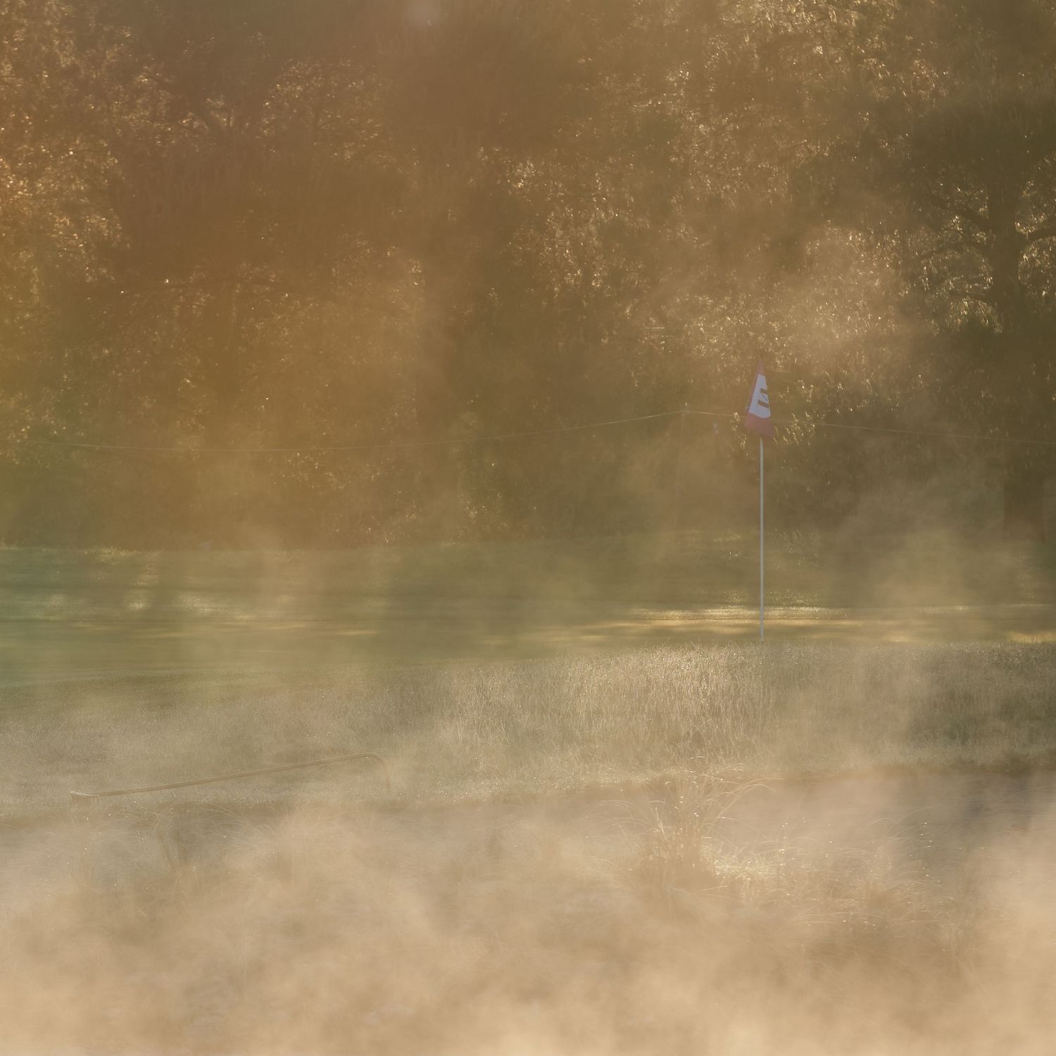 Misty morning golf course green with a flag pin barely visible through golden atmospheric fog.