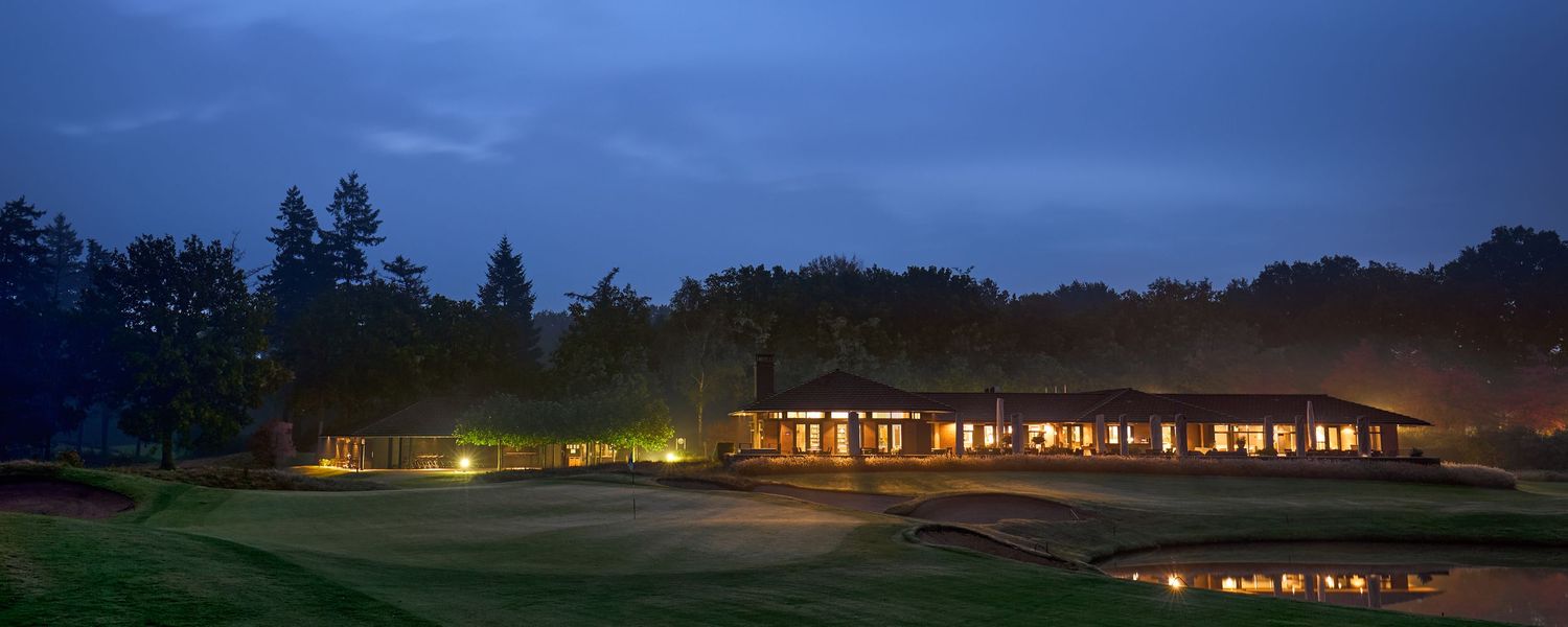 An illuminated golf clubhouse reflected in a pond at dusk, surrounded by misty trees and manicured greens.