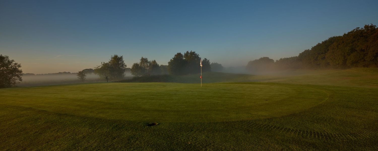 Peaceful misty morning on a golf course fairway with a flag pin and rolling hills in soft fog.
