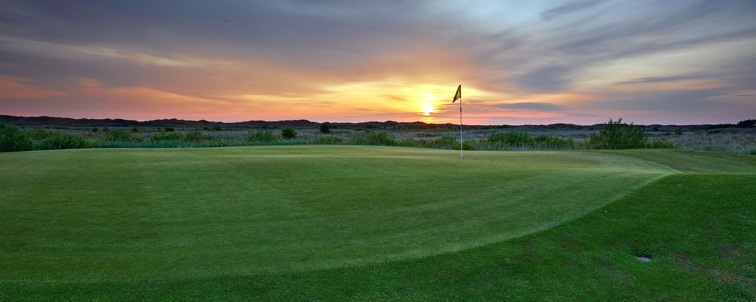 A golf course green at sunset with a flag pin silhouetted against a vivid dramatic sky.