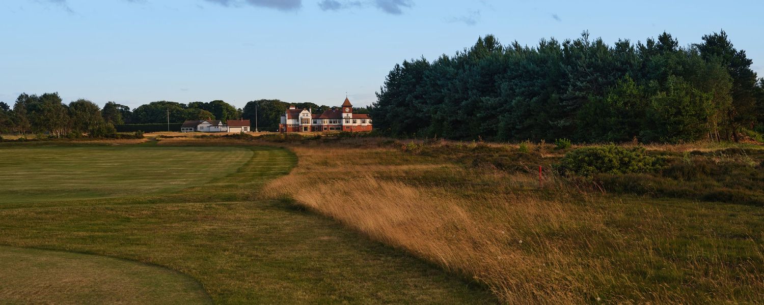 Expansive links golf course fairway at dusk with clubhouse and dense tree line in background.