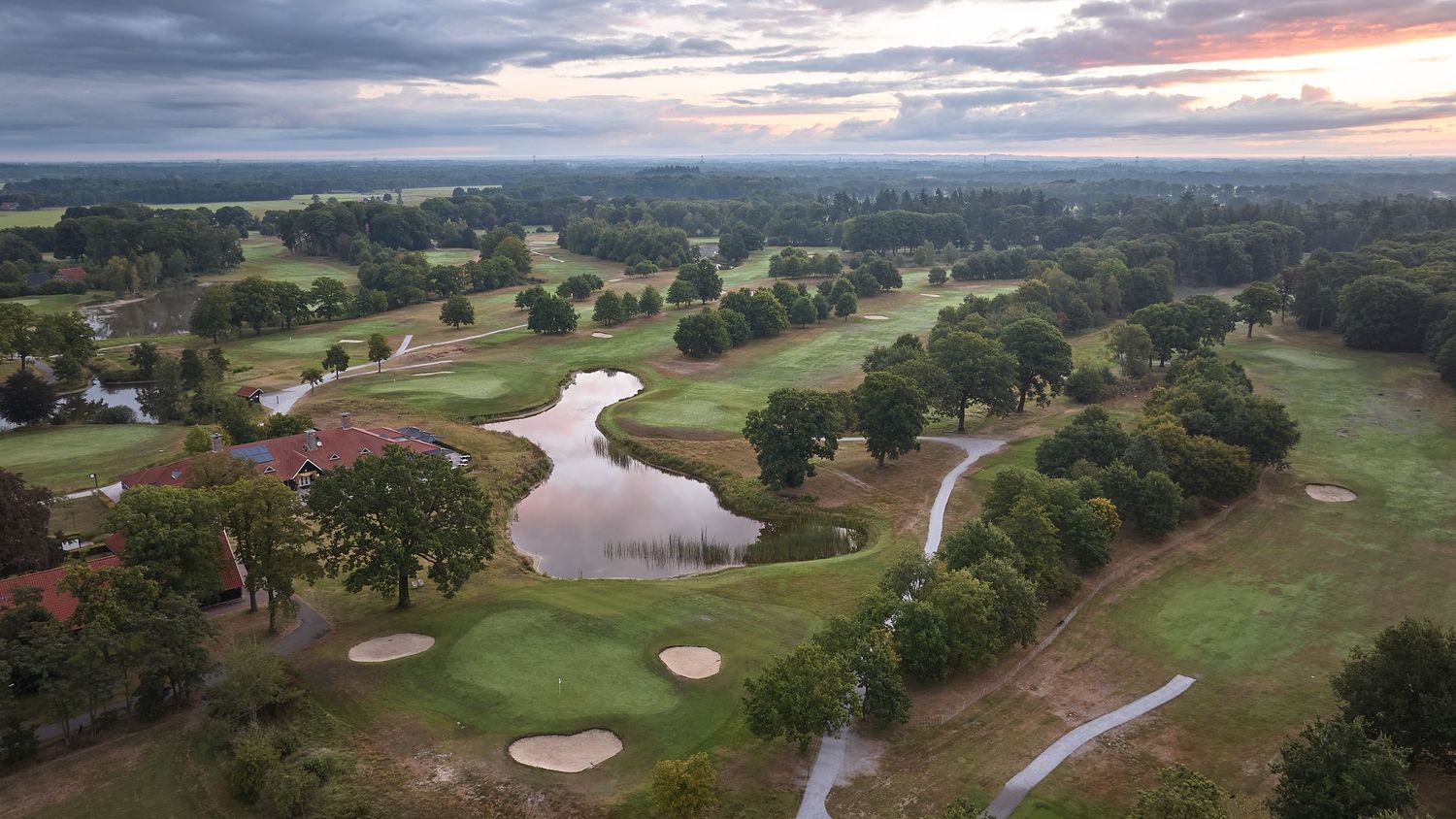 Aerial sunset view of a sprawling golf course with a winding pond, mature trees, and a red clubhouse building.