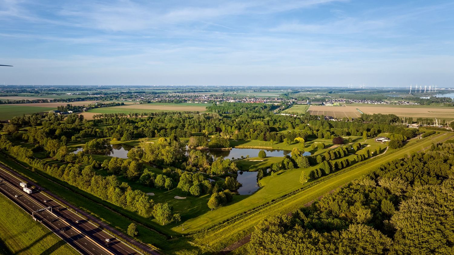 Aerial view of a golf course with ponds, trees, and a highway alongside open countryside.