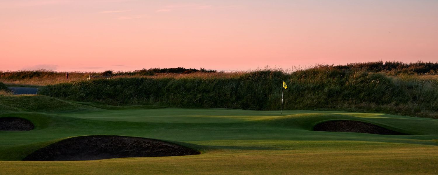 Links golf course at sunset with yellow flag, sand bunkers, and rolling green fairway under a pink and blue sky.