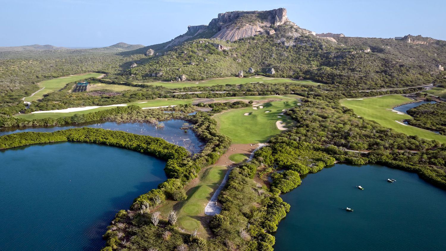 Aerial view of a golf course with lakes and lush greenery set against a dramatic rocky mountain.