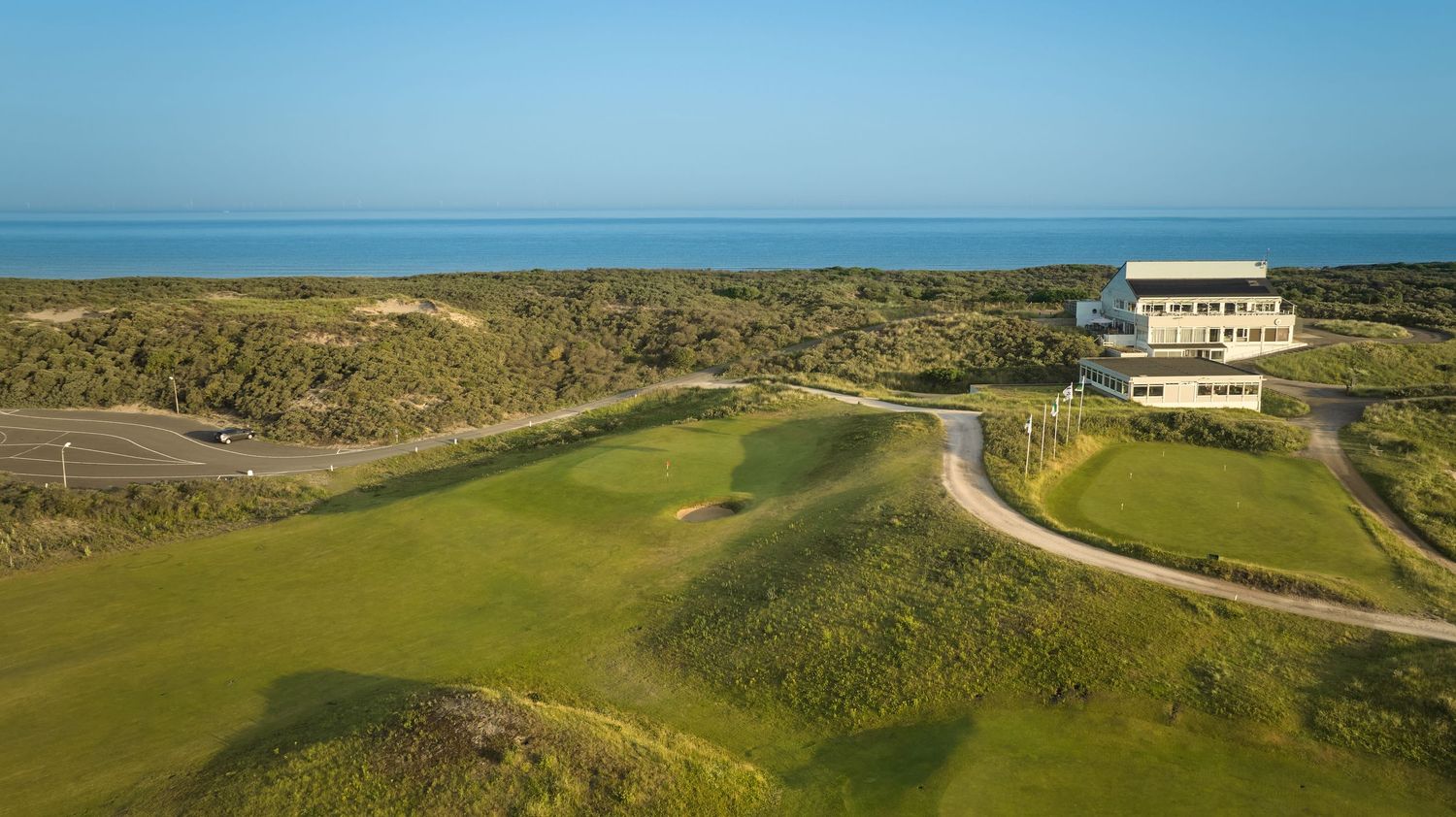 Aerial view of a coastal links golf course with a modern clubhouse, dunes, and the ocean in the background.
