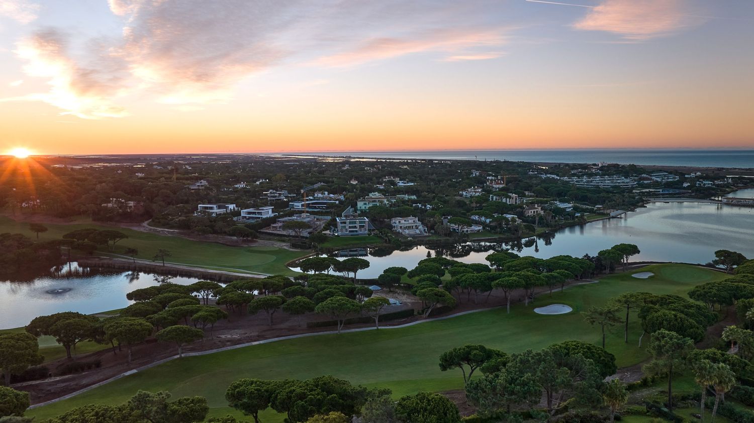 Aerial sunset view of a coastal golf course with water features, lush trees, and ocean horizon.