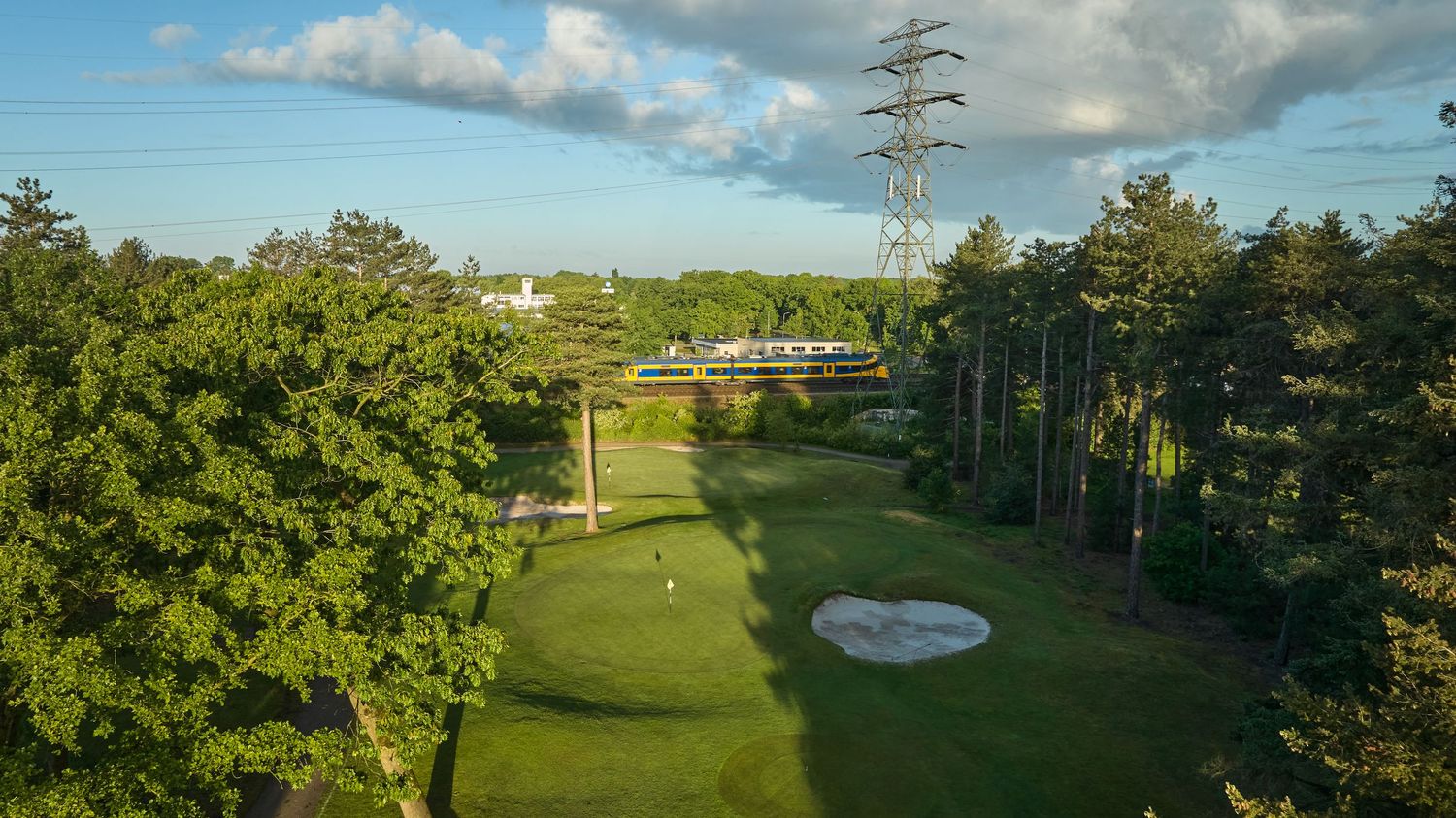 Aerial view of a golf course hole with sand bunker near a railway line and trees.