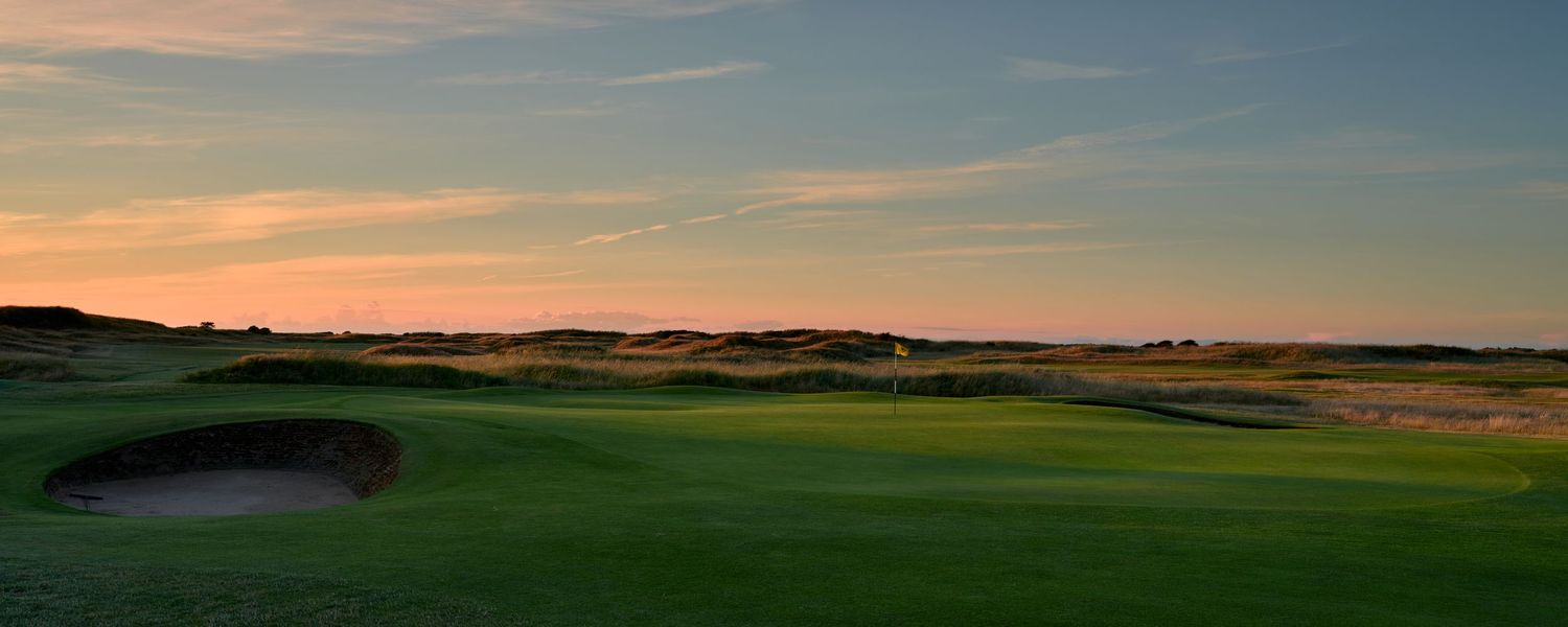 Panoramic view of links golf course at sunset with sand bunker, undulating fairway, and warm golden sky.