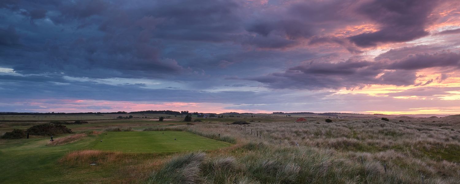 Panoramic links golf course at sunset with dramatic pink and purple clouds over vast open grassland terrain.