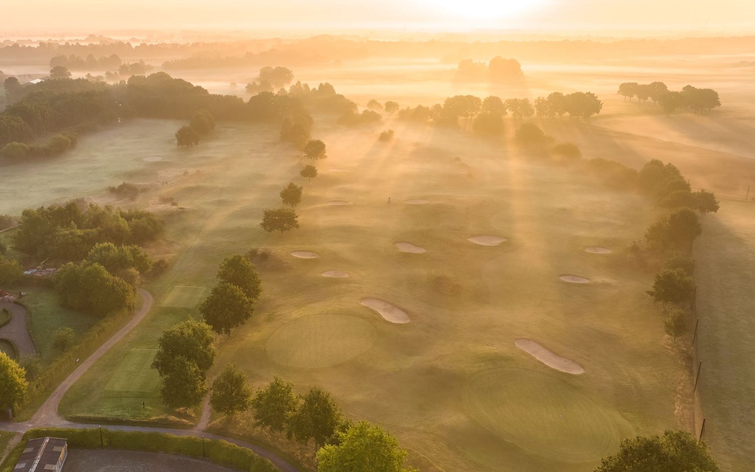 Aerial view of a misty golf course at sunrise with golden rays illuminating the fairways and bunkers.