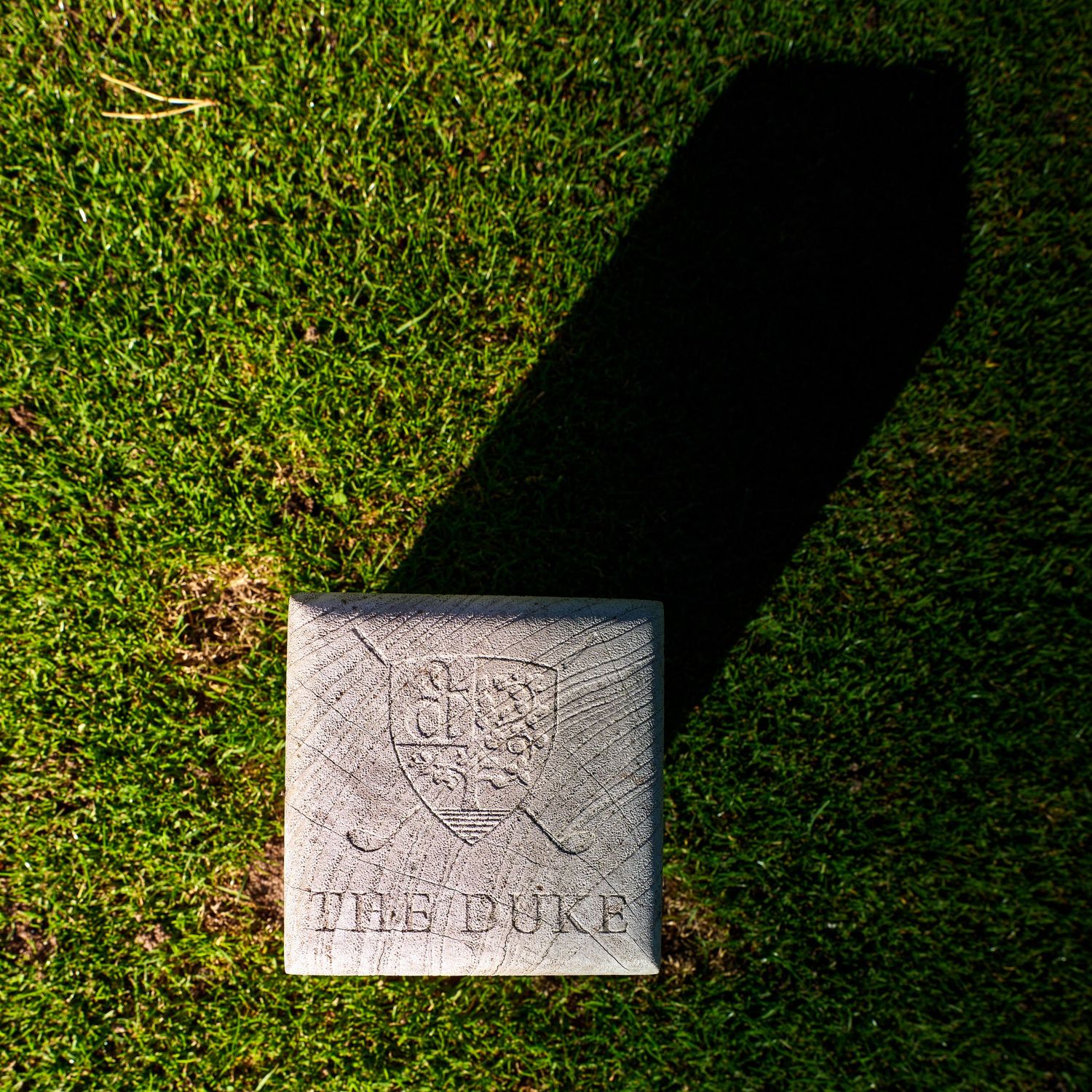 Stone marker engraved with The Duke golf club crest and crossed clubs on a manicured grass surface.