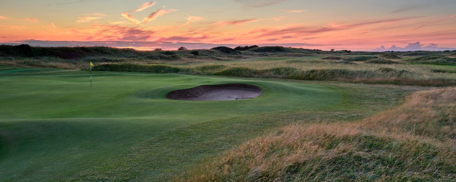 Links golf course at dusk featuring a deep bunker, wild rough, and a vivid pink and purple sky.
