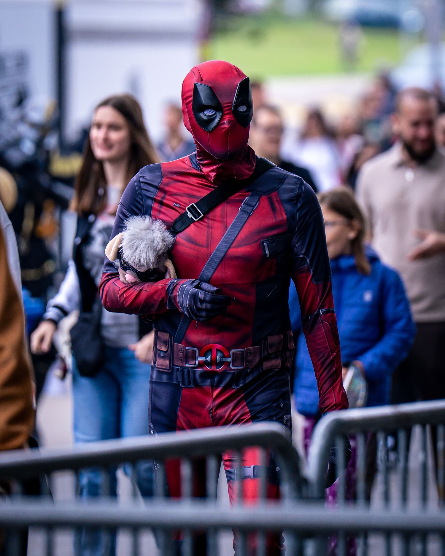 Person dressed in Deadpool costume holding a small fluffy toy at an outdoor comic convention event.