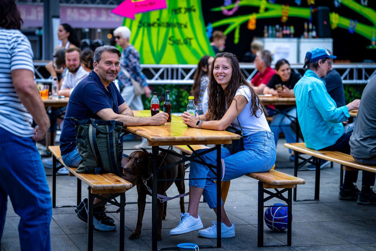 People enjoying outdoor dining at wooden tables with a dog present at a lively street event.