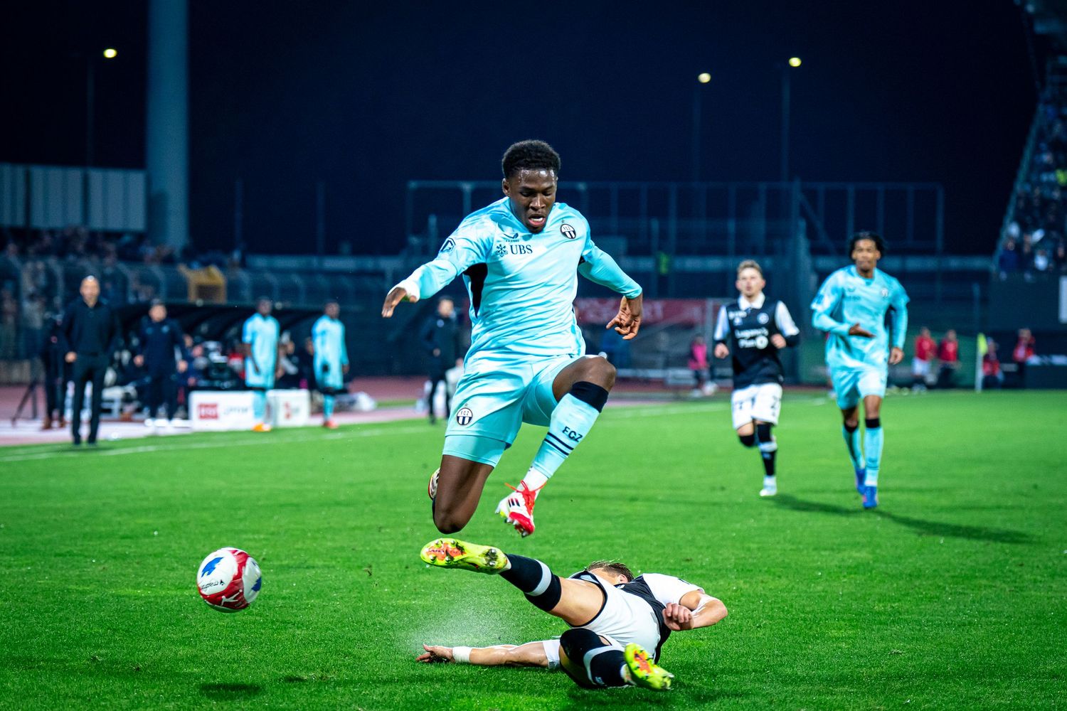 Soccer player in light blue kit jumping over sliding tackle during night match.