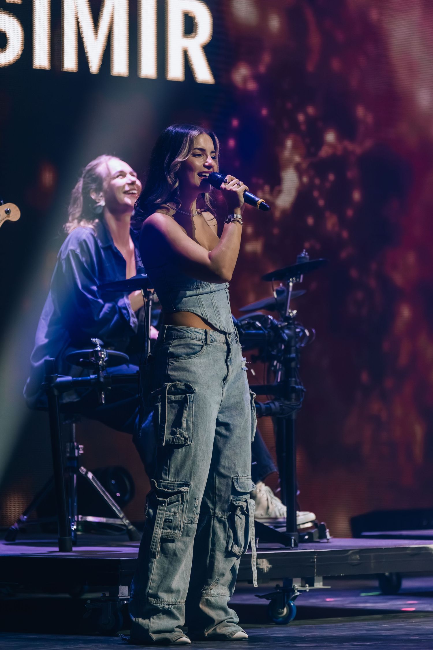 Female singer performing on stage with microphone, wearing cargo jeans, with band members and dramatic lighting behind her.