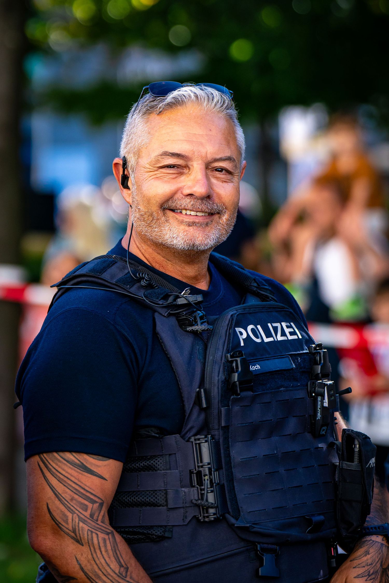 Smiling German Polizei officer in tactical vest with gray hair and tattoos posing outdoors at public event.