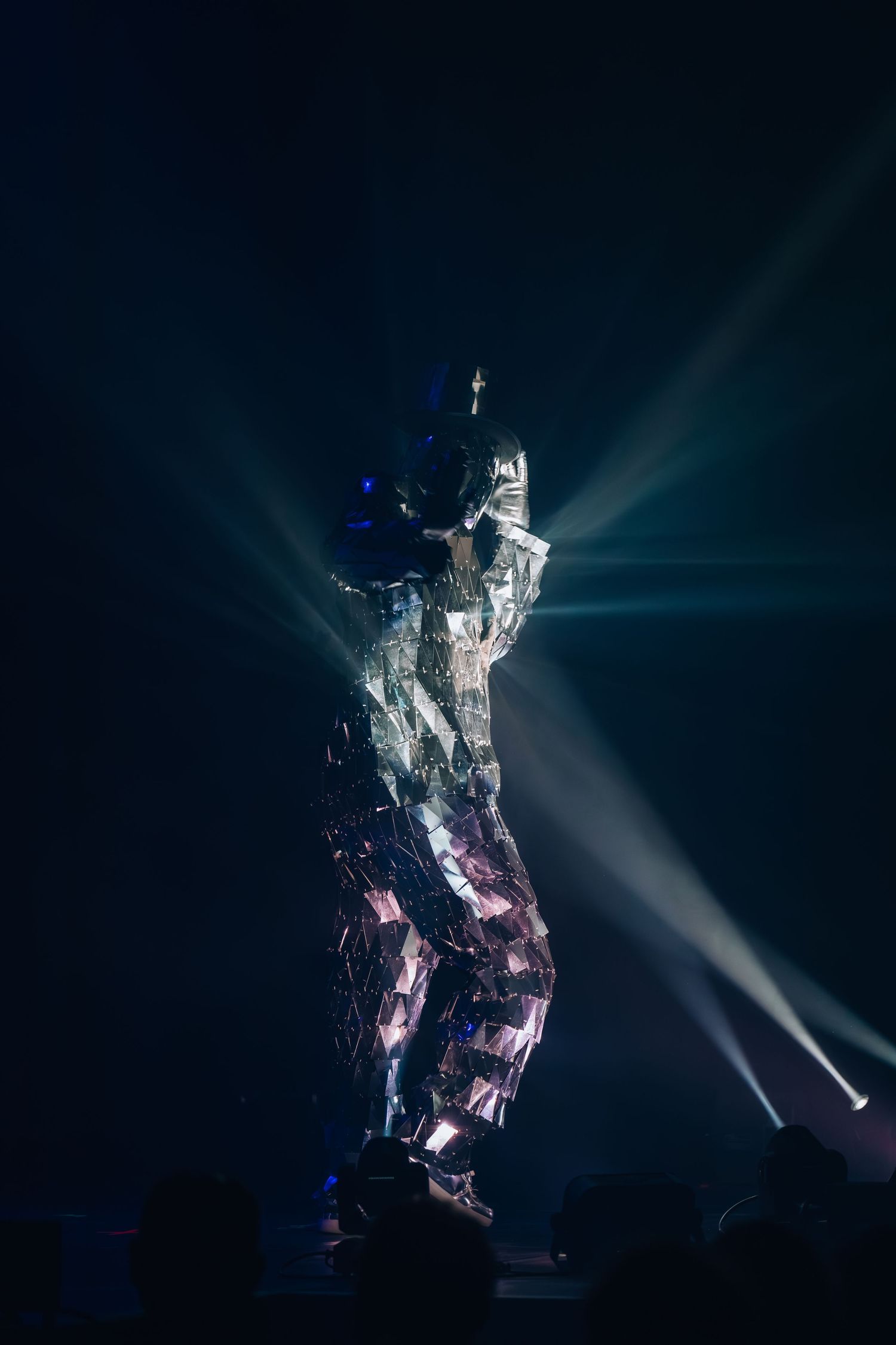 Performer in a reflective mirror costume dancing on stage under dramatic spotlights in a dark venue.