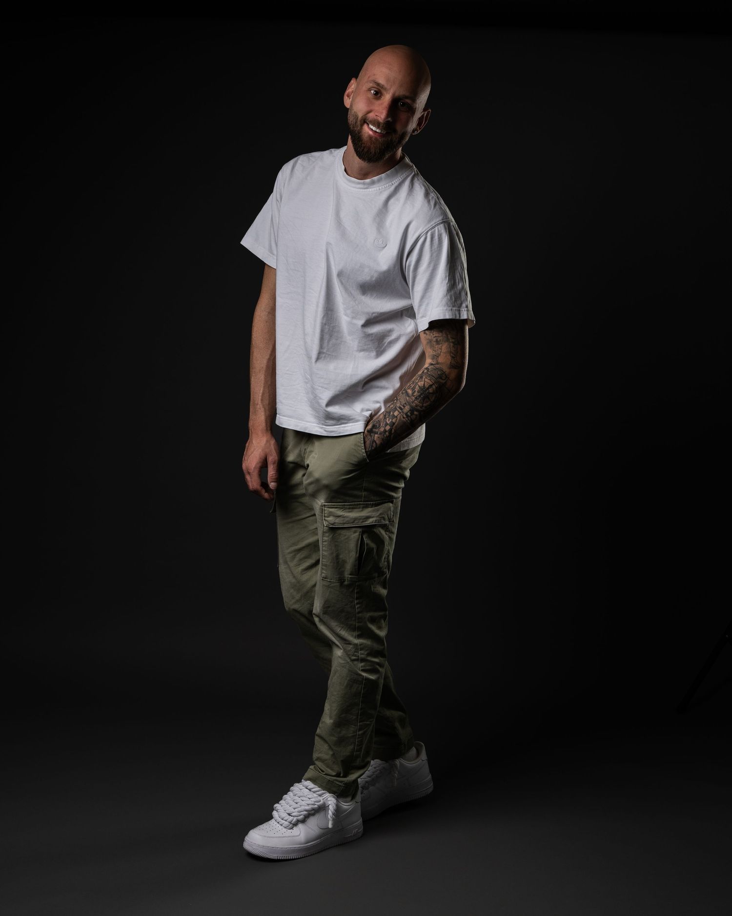 Bald tattooed man in white t-shirt and olive cargo pants posing against dark studio background.