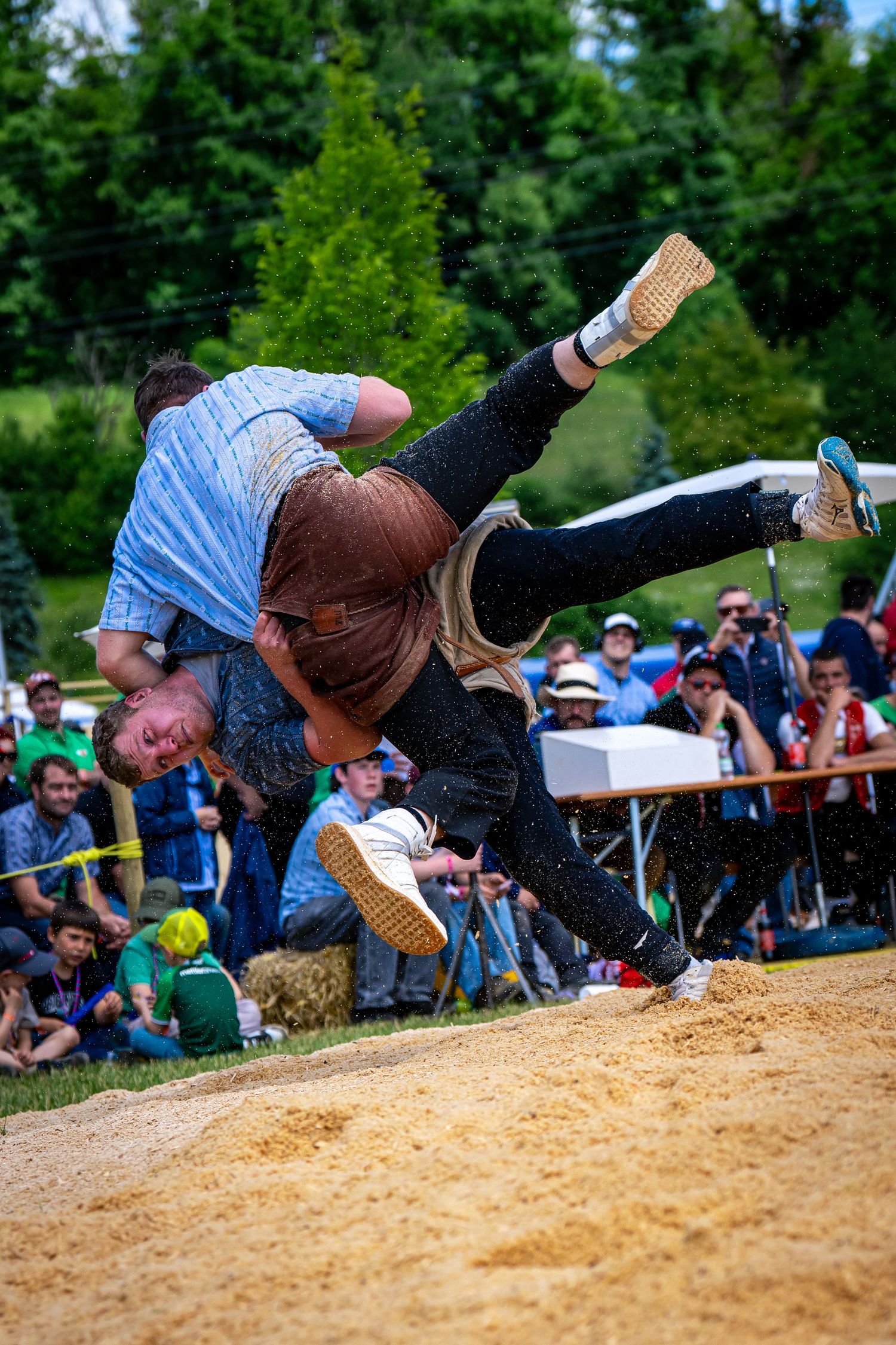 Two athletes compete in Swiss wrestling Schwingen on a sand pit, watched by a crowd of spectators outdoors.