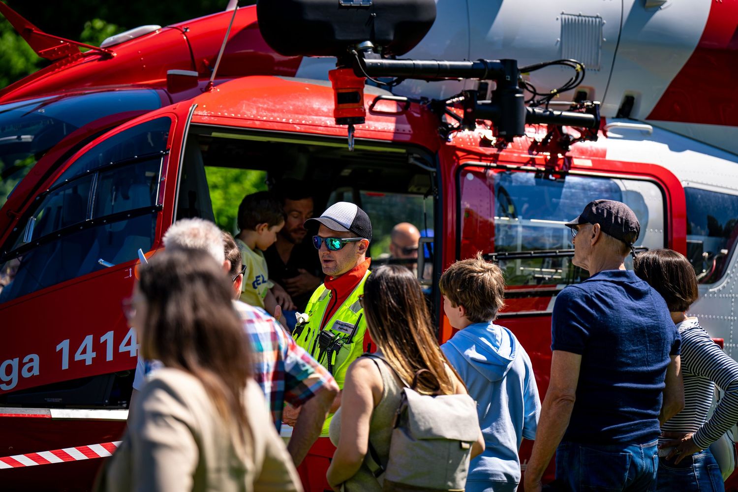 Crowd gathers around a red rescue helicopter numbered 1414 during a public demonstration on a sunny day.
