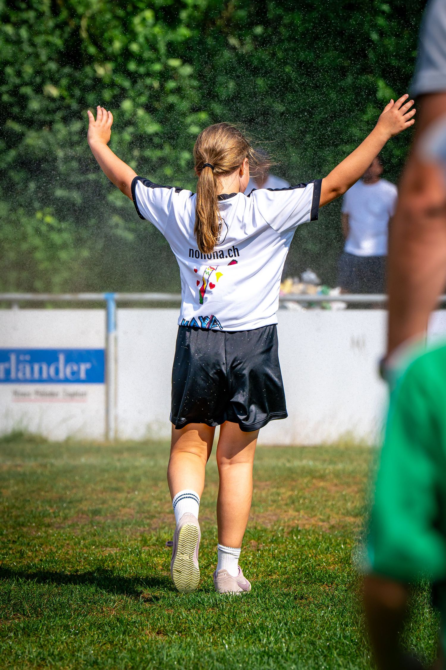 Young girl in white jersey raises arms in celebration on a green soccer field during a sunny outdoor match.