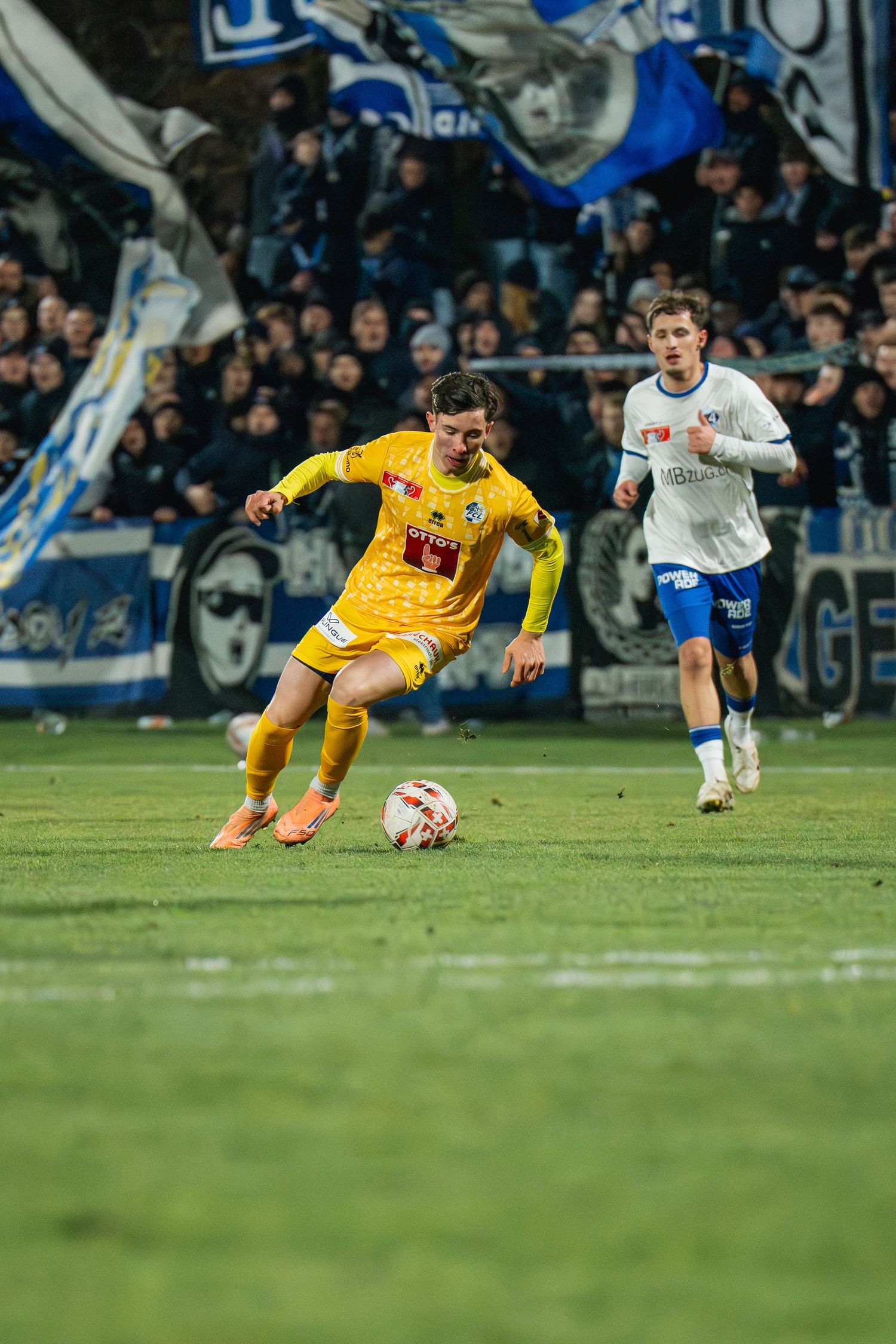Soccer player in yellow jersey dribbling ball during night match with fans waving blue and white flags in background.