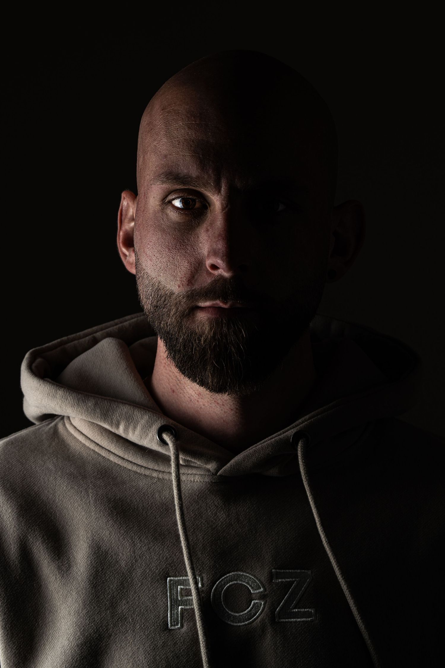Bald bearded man in dark FCZ hoodie, dramatic low-key portrait with moody lighting against black background.