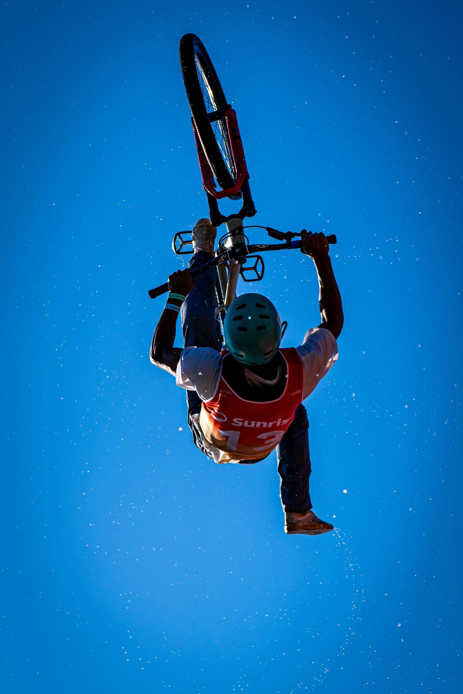 BMX rider performs an aerial trick against a clear blue sky, holding bike overhead while wearing a red jersey and helmet.