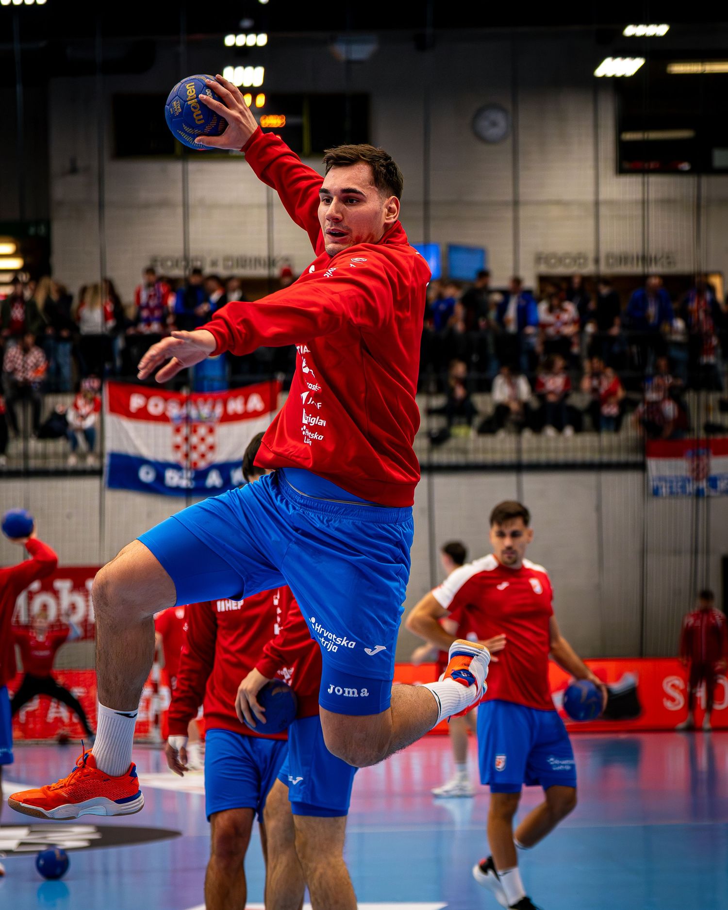 Croatian handball player jumps to throw ball during indoor match, wearing red jersey and blue shorts.