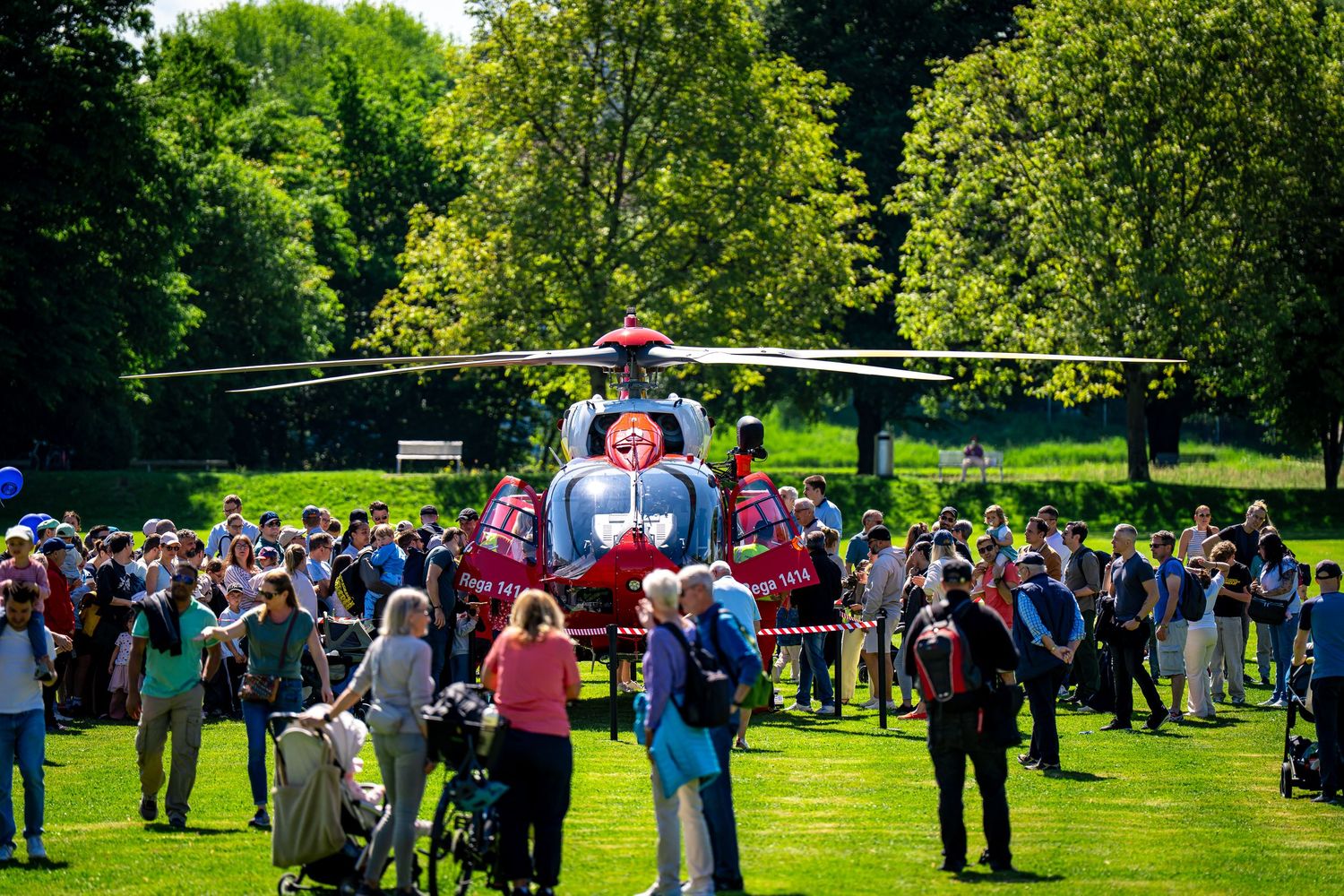 Crowd gathers around a red helicopter on green parkland on a sunny day with tall trees in the background.