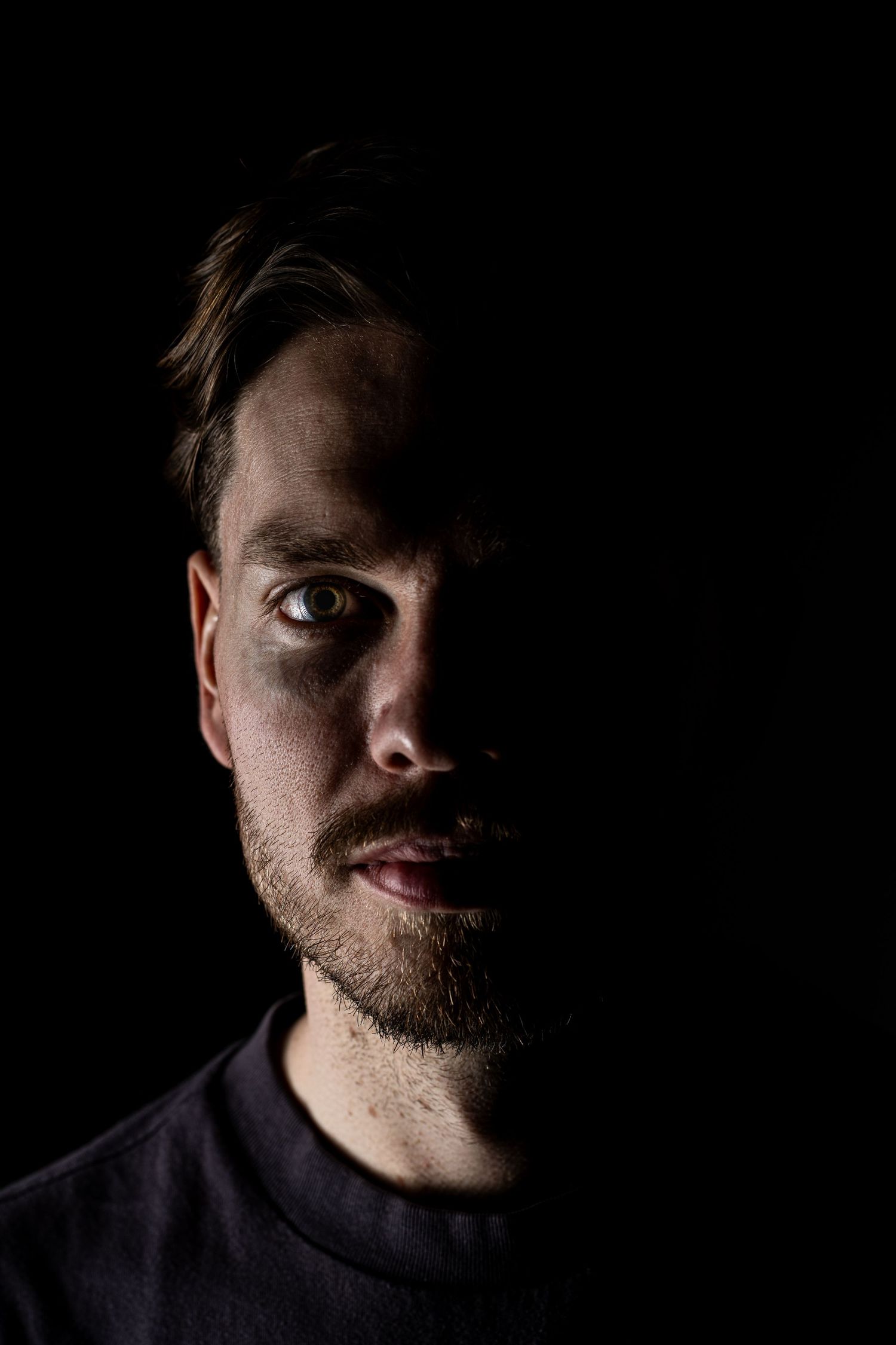 A dramatic low-key portrait of a young bearded man with half his face illuminated against a dark black background.