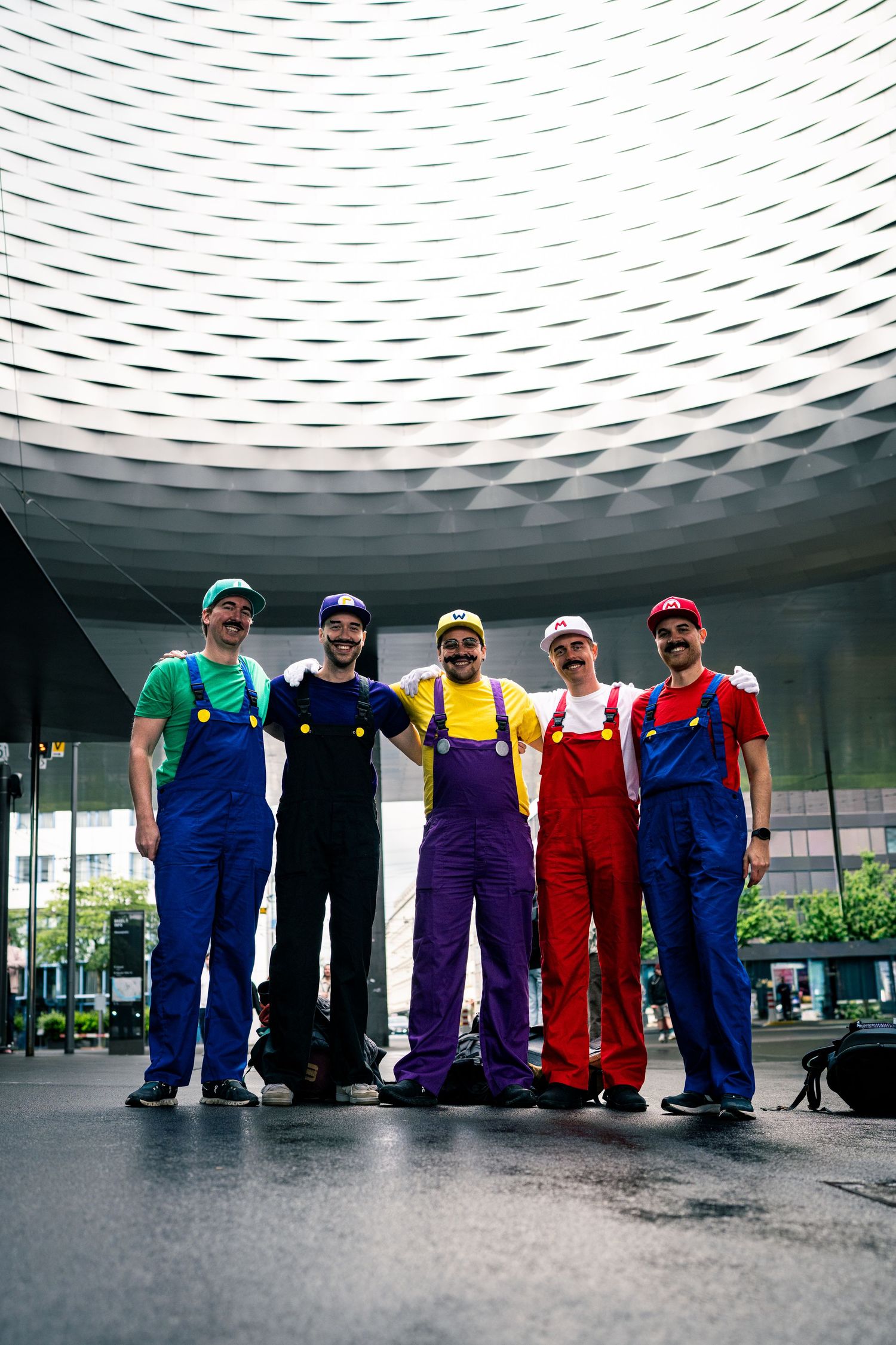 Five men in Mario Bros costumes pose together outside a modern building with a geometric ceiling.