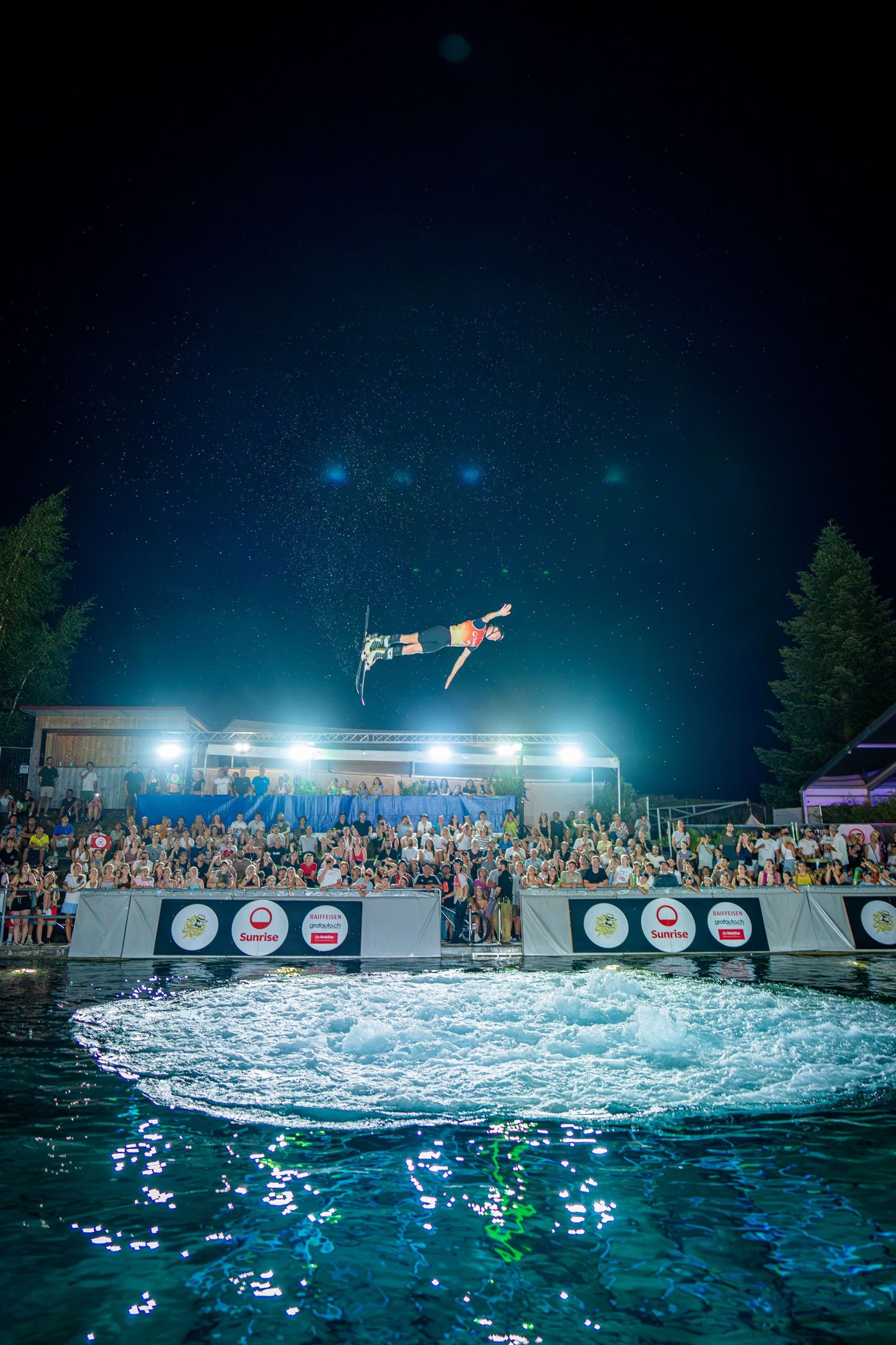 Diver soaring high above a illuminated pool at night during a spectacular cliff diving competition event.