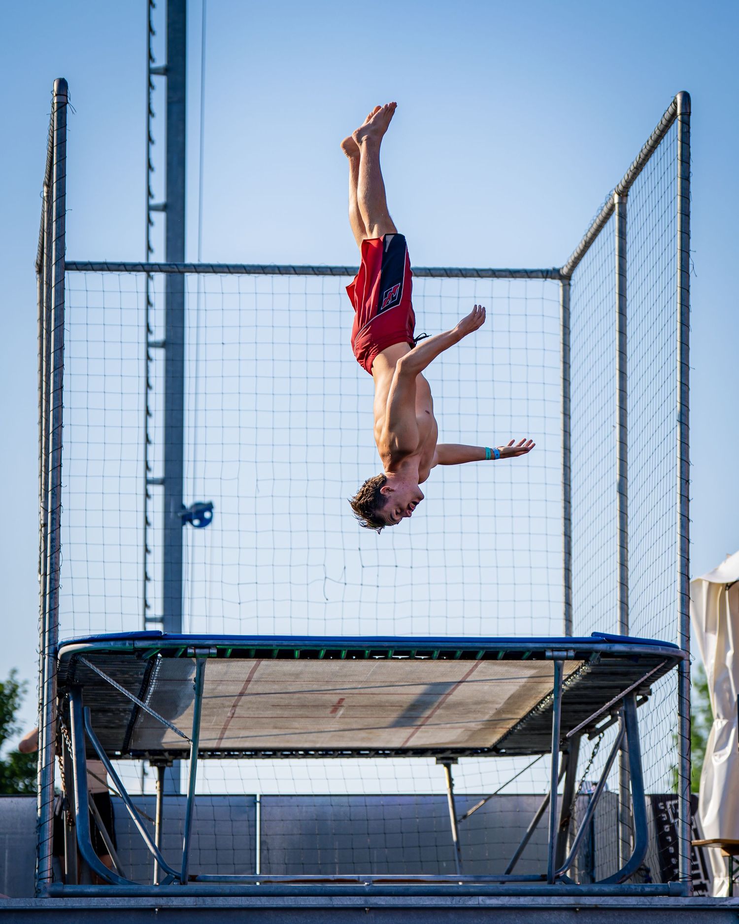 A shirtless boy performs a backflip on an outdoor trampoline against a clear blue sky.
