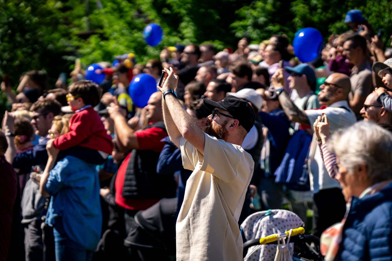 Large crowd gathered outdoors holding blue balloons, with a man in a white cap raising his hand at a sunny event.