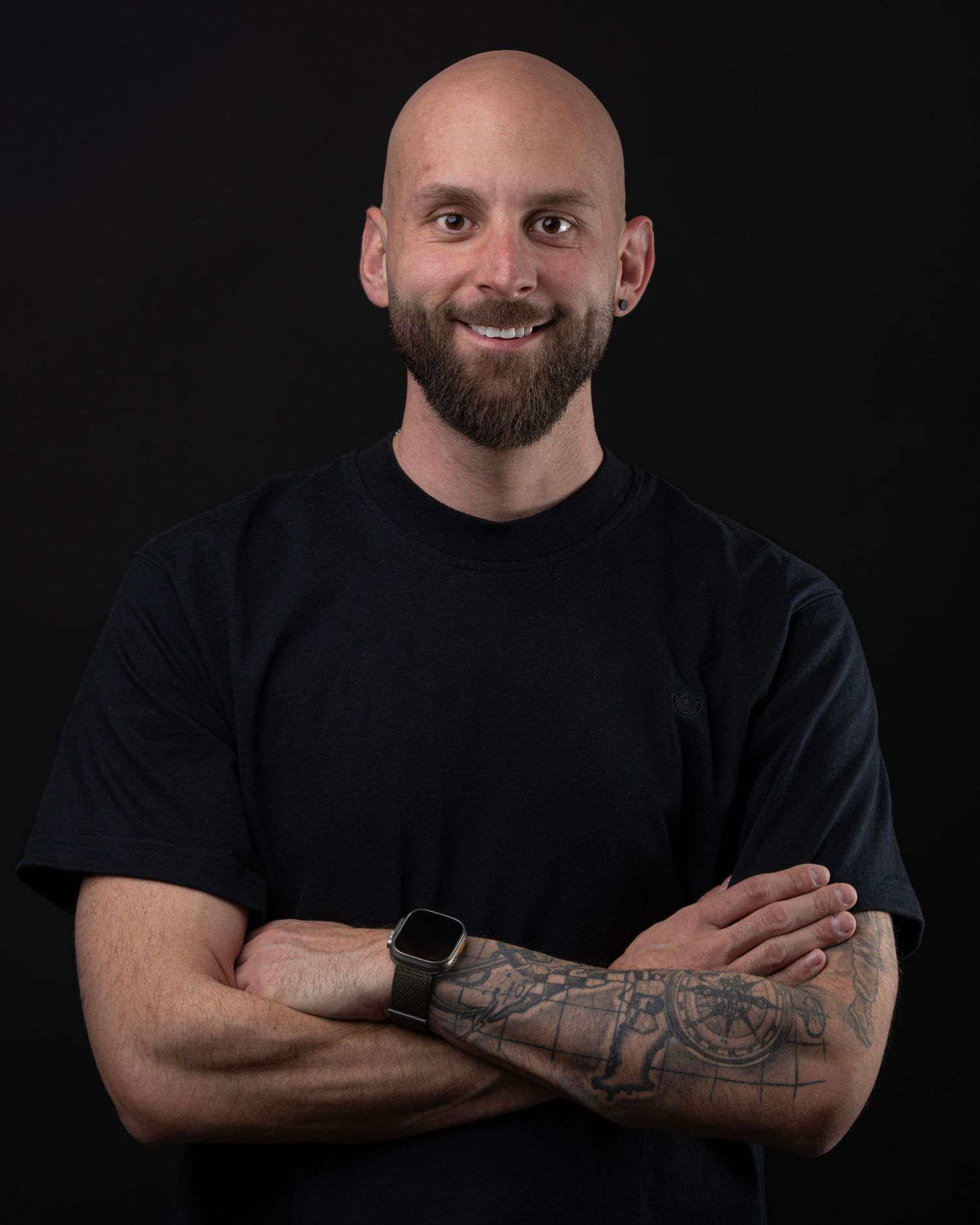 Bald man with beard and tattoos, arms crossed, wearing black t-shirt and smartwatch, smiling against dark background.