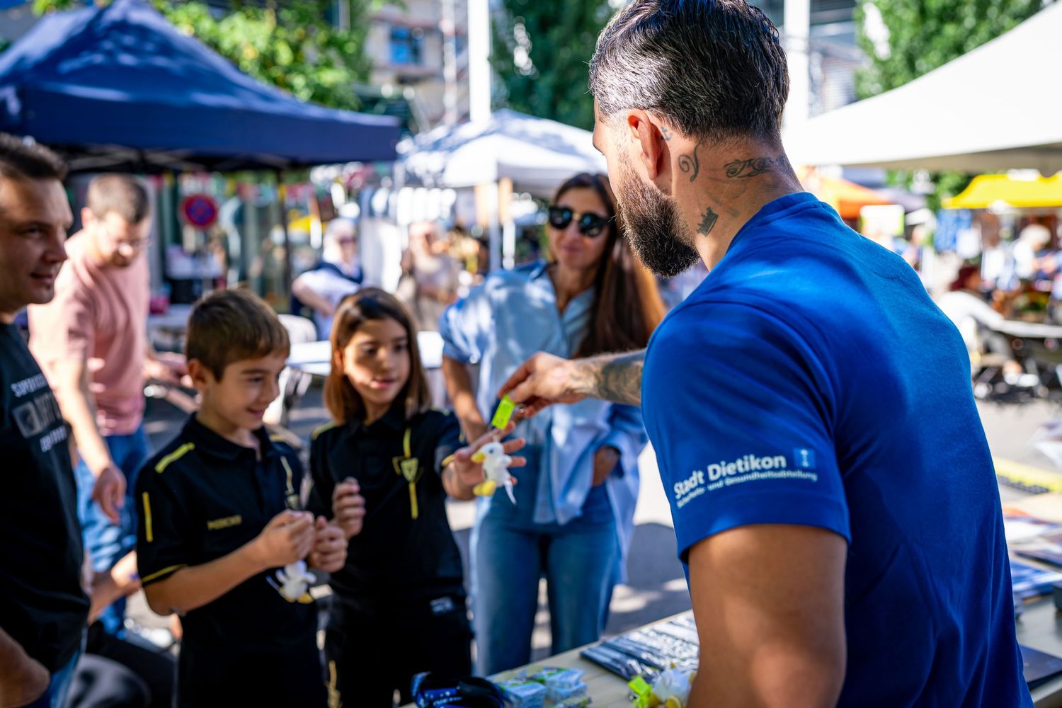 Man in blue shirt engages children at outdoor market booth on sunny day.