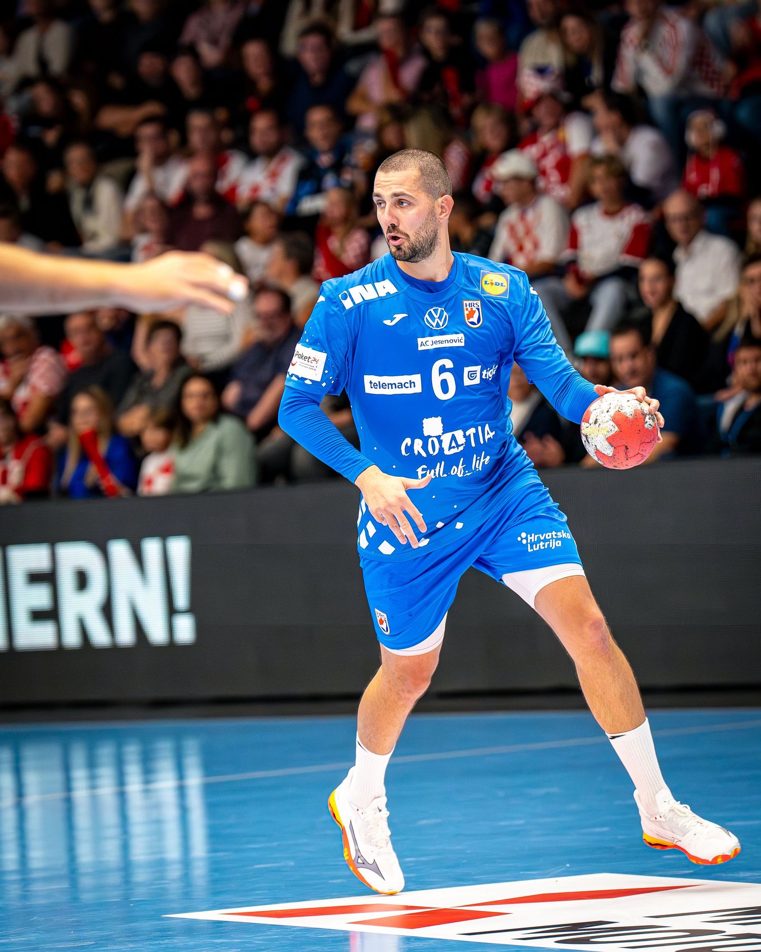 Croatia handball player number 6 in blue uniform holds ball during competitive indoor match with crowd in background.