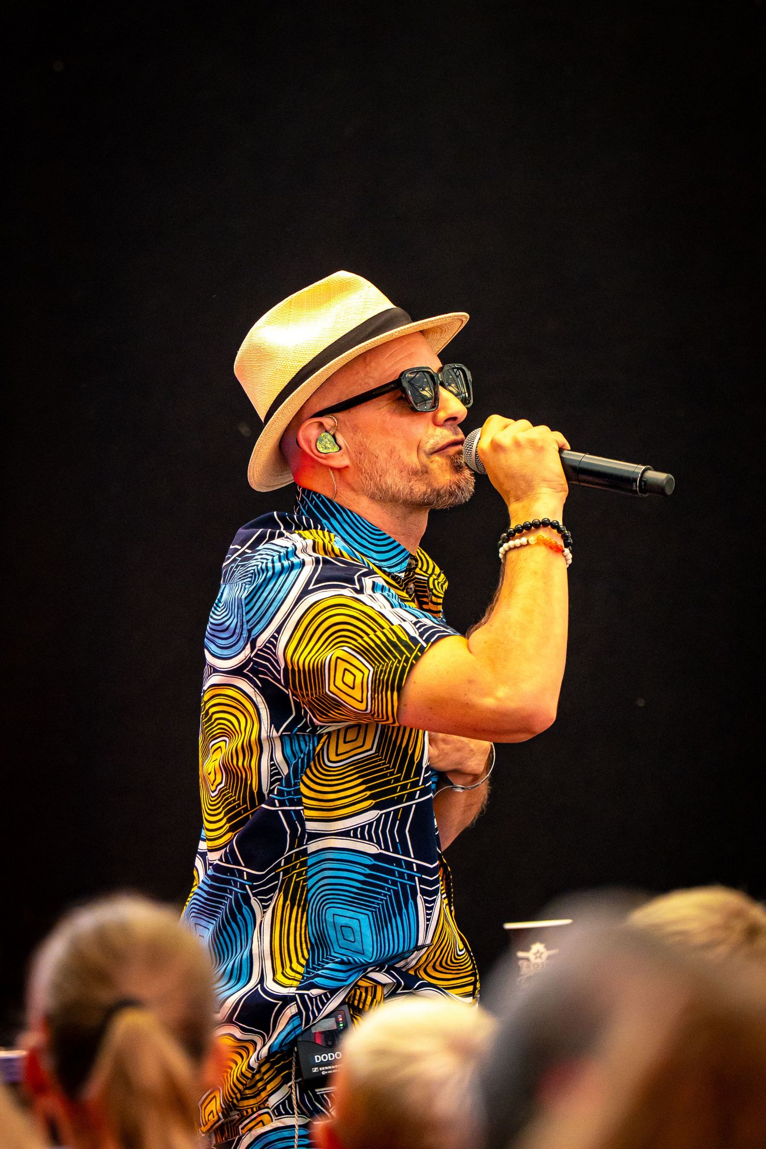 Male singer performing on stage wearing colorful patterned jumpsuit, straw fedora hat, and sunglasses while holding microphone.